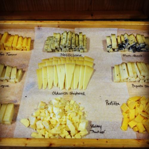 A wooden board displaying various cheese samples, labeled with names like Nettlesome, Oldwick Shepherd, and Valley Thunder.