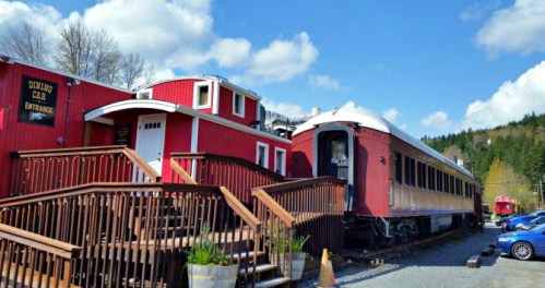 A vintage train car converted into a dining area, with wooden steps leading up to it, surrounded by trees and blue skies.