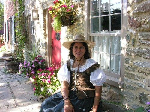 A woman in a historical costume sits outside a stone cottage, surrounded by flowers and greenery.