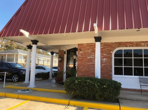 Exterior of a building with a red metal roof, brick walls, white columns, and a landscaped entrance.