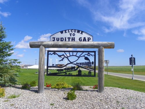 Welcome sign for Judith Gap, featuring a scenic design with mountains and wildlife, set against a blue sky.