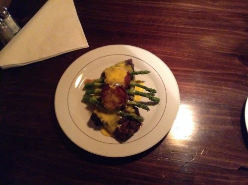 A plate of steak topped with melted cheese, served on a bed of asparagus and a side of potato.