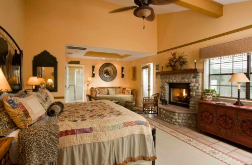 Cozy bedroom with a quilted bed, stone fireplace, and warm yellow walls, featuring a seating area and decorative accents.
