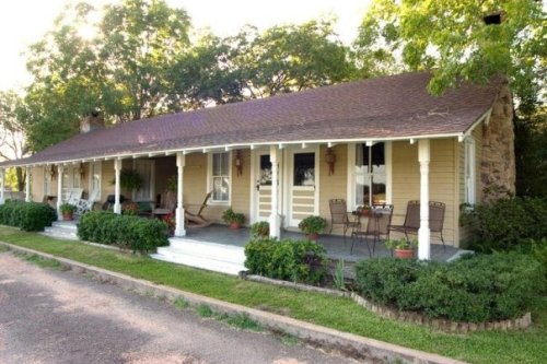 A charming single-story house with a porch, surrounded by greenery and potted plants, on a sunny day.