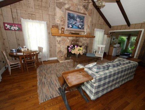 Cozy living room with wooden walls, a stone fireplace, a plaid couch, and a small dining table by the window.
