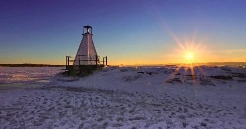 A snowy landscape at sunset, featuring a lighthouse silhouette and rays of sunlight illuminating the scene.