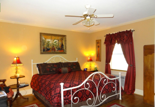 Cozy bedroom with a decorative bed, warm lighting, and rich red curtains, featuring a ceiling fan and artwork on the walls.
