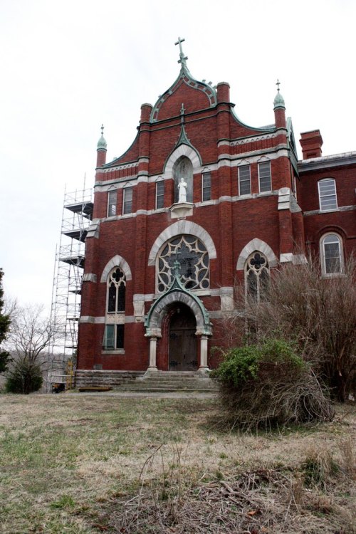 Historic brick church with a statue, scaffolding, and overgrown grass in front, under a cloudy sky.