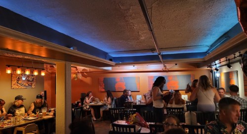A lively restaurant scene with diners at tables, colorful lighting, and wall art depicting maps.