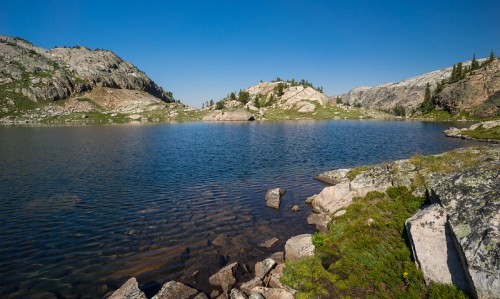 A serene lake surrounded by rocky hills and greenery under a clear blue sky.