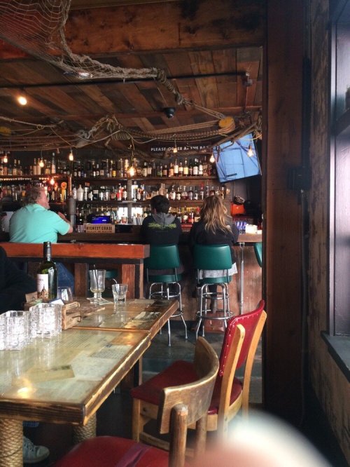 A cozy bar scene with patrons sitting at the counter, surrounded by shelves of bottles and rustic wooden decor.