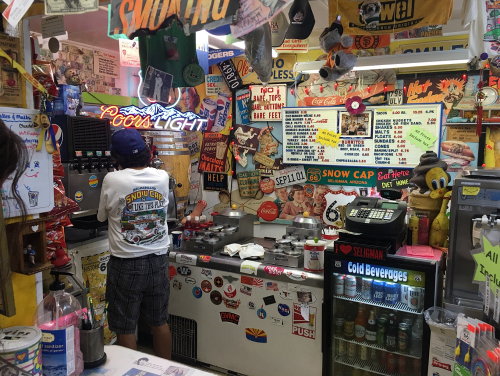 A colorful convenience store interior filled with signs, stickers, and a person preparing drinks at the counter.
