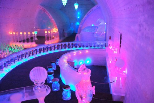 A colorful ice bar with illuminated ice sculptures, glowing lights, and a curved bar surrounded by ice seating.