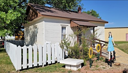 A small white house with a brown roof, surrounded by a white picket fence and decorated with flowers and a figure in a blue dress.
