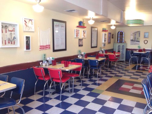 A retro diner interior with checkered floors, red and blue chairs, and vintage decor on the walls.