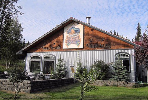 A rustic building with a sign reading "St. Elias Brewing Company," surrounded by greenery and outdoor seating.