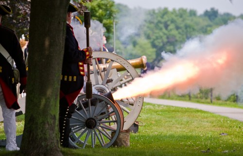 A historic cannon fires, releasing smoke and flames, with soldiers in period uniforms standing nearby.