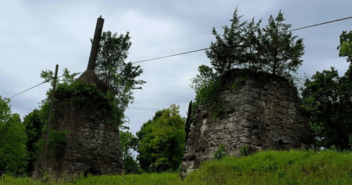 Two weathered stone structures overgrown with greenery, set against a cloudy sky.