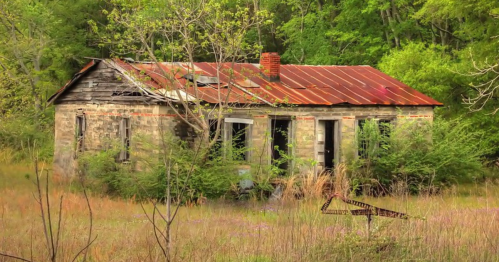 Abandoned stone house with a rusty roof, surrounded by overgrown grass and trees in a rural setting.