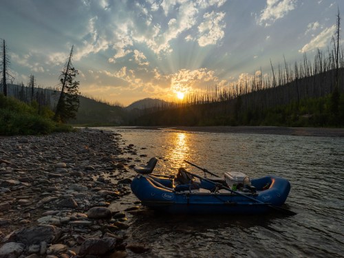 A blue raft rests on a rocky riverbank at sunset, surrounded by trees and mountains under a colorful sky.