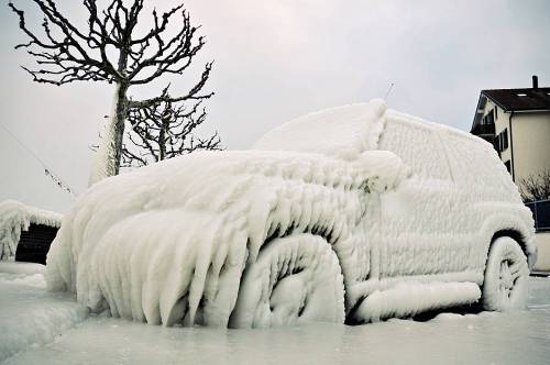 A car covered in thick layers of ice and snow, with frozen details and a wintery landscape in the background.