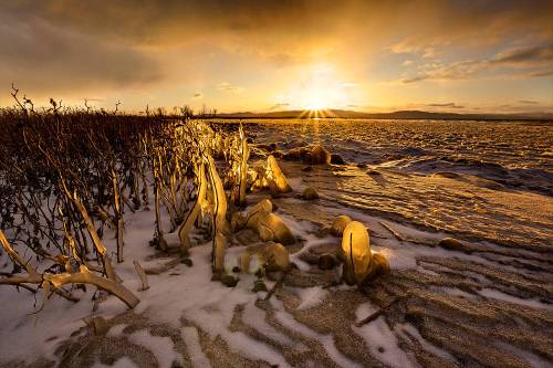 A winter landscape at sunset, featuring icy formations and a golden glow over a snowy shoreline.