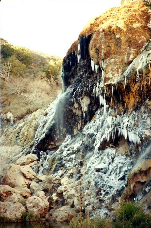 A cascading waterfall with icicles, surrounded by rocky terrain and sparse vegetation under a clear sky.