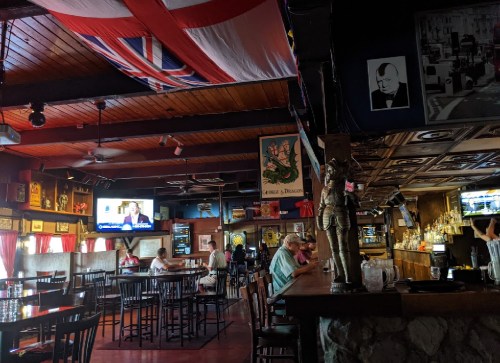 Interior of a pub with wooden decor, a large English flag, and patrons seated at tables and the bar.