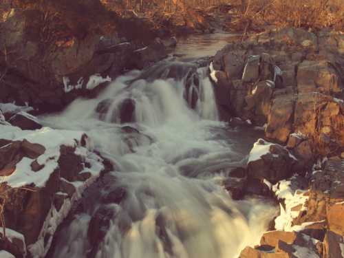 A serene waterfall cascades over rocky terrain, surrounded by bare trees and patches of snow.