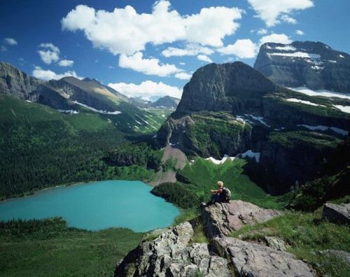 A person sits on a rocky ledge overlooking a turquoise lake surrounded by lush green mountains and a blue sky with clouds.