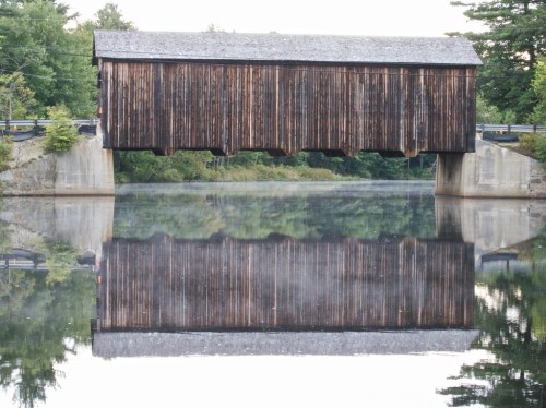 A wooden covered bridge spans a calm river, reflecting its image in the water below, surrounded by trees.