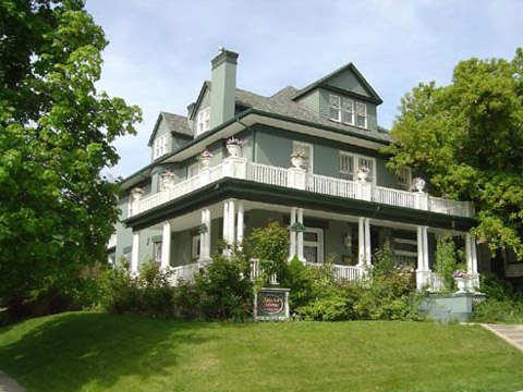 A large, green Victorian-style house with a wraparound porch and lush greenery surrounding it.