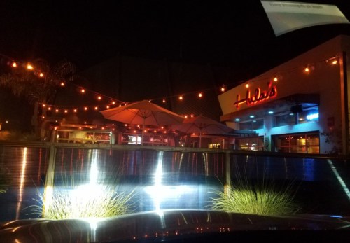 A nighttime view of a restaurant with string lights and umbrellas, reflecting in a water feature.