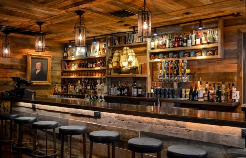 A rustic bar with wooden decor, shelves of liquor, and dim lighting, featuring high stools and a ship model display.