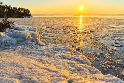 Sunset over a frozen lake, with ice formations and shimmering water reflecting warm golden hues.
