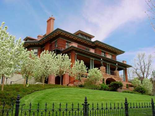 A large, elegant brick mansion surrounded by flowering trees and a green lawn, under a blue sky.