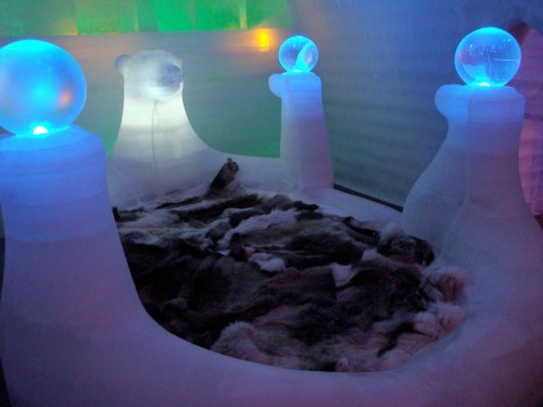 A cozy ice bed with fur blankets, illuminated by glowing blue orbs, set in a colorful ice room.