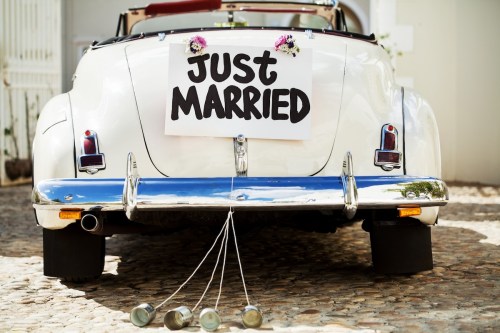 A white vintage car with a "Just Married" sign and cans tied to the back, parked on a cobblestone street.