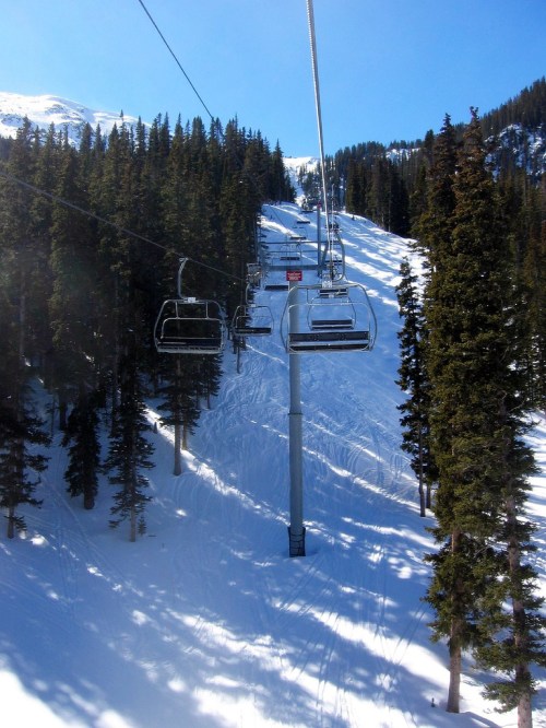 A ski lift ascends a snowy mountain surrounded by tall pine trees under a clear blue sky.