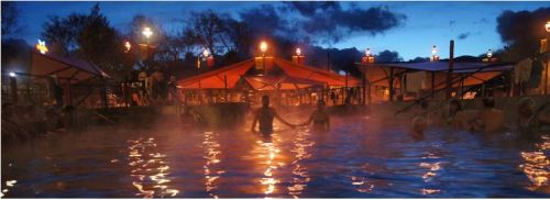 A serene hot spring at dusk, with people relaxing in the water and glowing lanterns illuminating the surroundings.