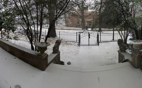 A snowy scene featuring a gated entrance, flanked by stone lion statues, with trees and a building in the background.