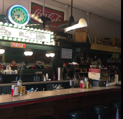 A vintage diner counter with a clock, Coca-Cola signage, and various condiments and drinks on display.