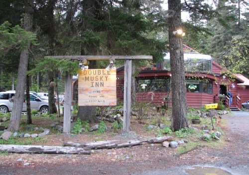 Sign for Double Musky Inn surrounded by trees, with a rustic building in the background and a gravel path.