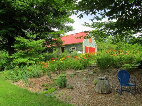 A charming house with a red roof surrounded by vibrant flowers and greenery, featuring a blue chair and a garden area.