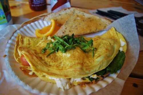 A fluffy omelette filled with spinach and tomatoes, served with toast and a slice of orange on a paper plate.