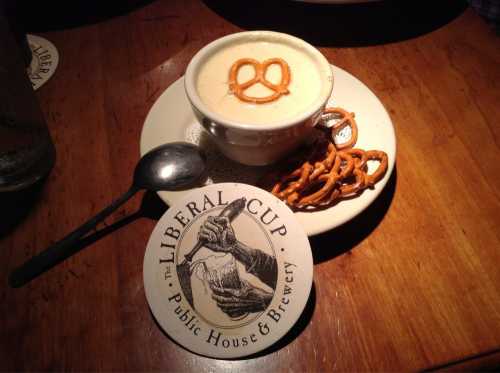 A bowl of creamy soup topped with a pretzel, accompanied by a coaster from Liberal Cup Brewery and a spoon.