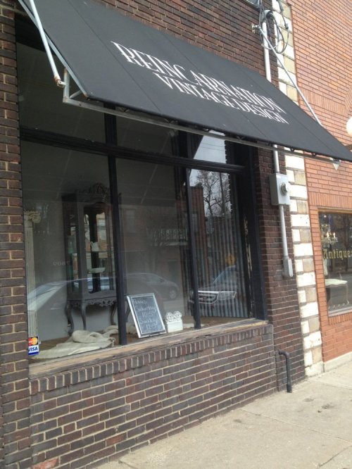 Exterior of a vintage shop with a black awning, large windows, and a brick facade. A sign is visible in the window.