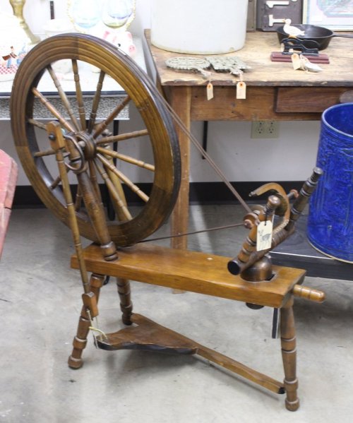 A vintage wooden spinning wheel with a large wheel and a foot pedal, displayed in a rustic setting.