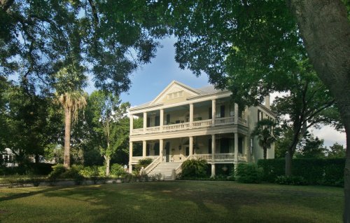 A large, historic house with a wraparound porch, surrounded by lush greenery and trees under a clear blue sky.