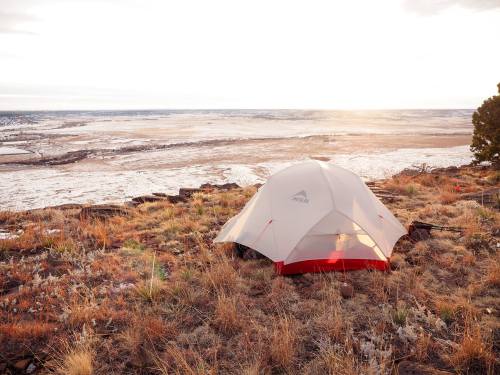 A tent set up on a rocky hillside, overlooking a vast, open landscape at sunset.